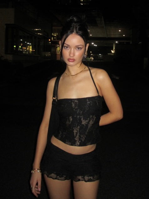 Model wearing a matching black lace set from Yasmin The Label featuring a halter top with adjustable ties at the top and back, and a mini skirt with built-in shorts; intricate lace fabric with triple-layered coverage on bust area, double-layered throughout; model is an Australian size 6 and wears XS/S in both pieces.