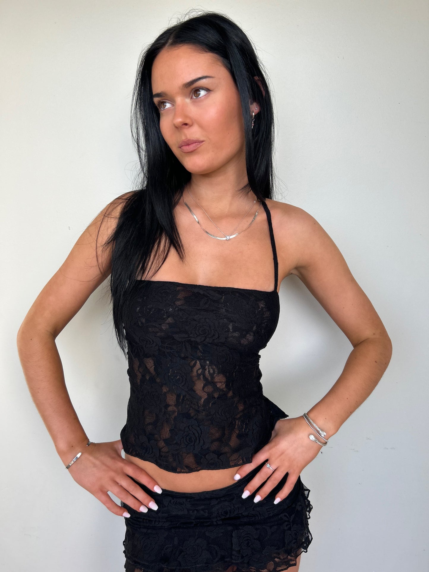 Model wearing a matching black lace set from Yasmin The Label featuring a top with adjustable ties and mini skirt with built-in shorts; triple-layered lace on bust area for coverage, double-layered throughout; model is size 6 and wears XS/S.