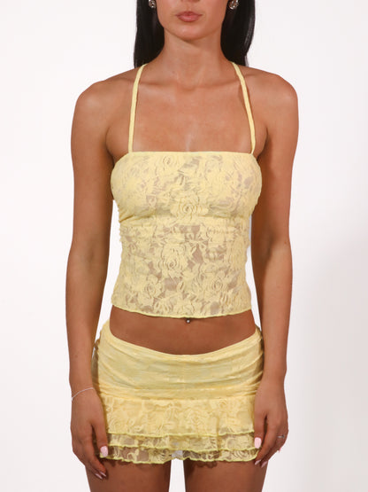 Model wearing a matching yellow lace set from Yasmin The Label featuring a top with adjustable ties and mini skirt with built-in shorts; triple-layered lace on bust, double-layered throughout; model is size 6 and wears XS/S.