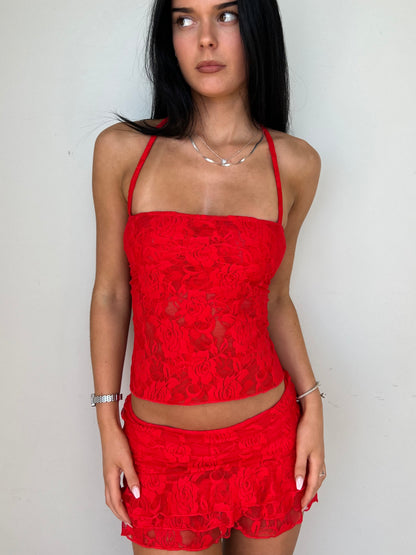Model wearing a matching red lace set from Yasmin The Label featuring a top with adjustable ties and mini skirt with built-in shorts; triple-layered lace on bust area for coverage, double-layered throughout; model is size 6 and wears XS/S.