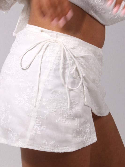 Model wearing a matching white set from Yasmin The Label featuring a scoop neck top with adjustable ties at the top and back, and a mini skirt with inbuilt shorts and embroidered pattern; skirt designed to sit low waisted with two ties at the side for adjustability; clean minimalist design shown on model who is an Australian size 8 and wears S/M in both pieces.